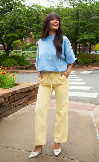 Woman wearing a light blue sweater and yellow pants standing on a sidewalk with trees and flowers in the background.