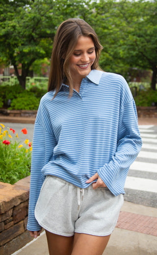 Woman wearing a blue striped sweater and gray shorts standing outdoors with greenery in the background