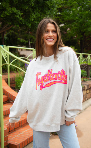 Woman wearing a sweatshirt with 'Fayetteville' printed on it, standing outdoors.