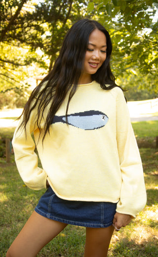 A woman wearing a pale yellow crewneck sweater with a pixelated fish graphic on the front, paired with a denim mini skirt, standing outdoors in a grassy park area with trees in the background.