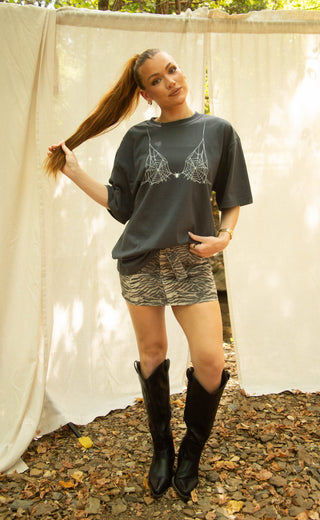A woman stands outdoors in front of draped white fabric, wearing an oversized dark gray graphic T-shirt with a spiderweb design, patterned mini shorts, and black cowboy boots.