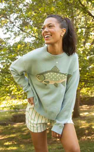 Woman wearing blue crewneck sweater with long sleeves and knit green fish. 
