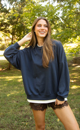 A woman wearing an oversized navy blue crewneck sweatshirt with a white undershirt peeking out at the hem, paired with black lace shorts, standing outdoors in a grassy park with trees in the background.