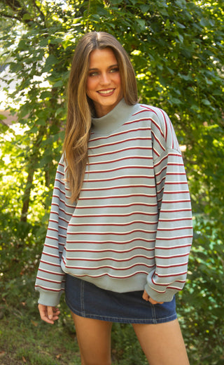 A woman wearing a light gray turtleneck sweater with thin red and navy horizontal stripes, paired with a blue denim mini skirt, standing outdoors in front of a lush green background.