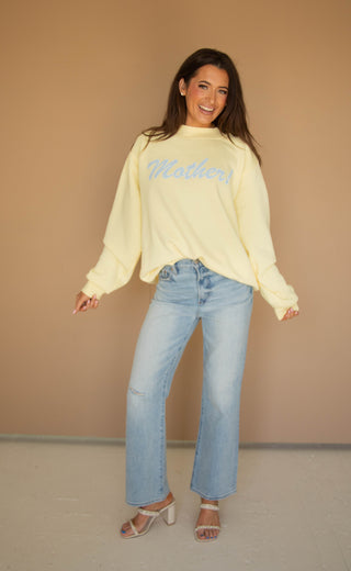 Woman wearing a light yellow mockneck sweatshirt with blue "mother!" text and blue jeans against a beige background