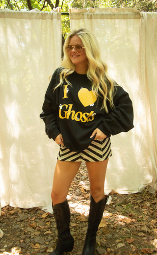 A woman wearing a black crewneck sweatshirt with a yellow 'I Heart Ghost' graphic, paired with black and white striped shorts and black cowboy boots, posing outdoors in front of draped white fabric backdrops.