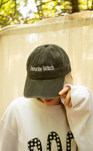 A person outdoors wearing a dark olive green baseball cap with 'Favorite Witch' embroidered in white on the front, paired with a white sweatshirt.