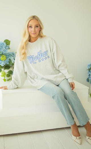 Woman wearing an ash grey mockneck sweatshirt with 'Mother' text in blue, sitting on a white couch with flowers in the background.