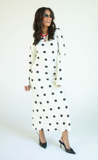 A woman modeling a long-sleeve midi dress with a black polka dot pattern on a cream background, styled with black pointed-toe heels and sunglasses against a plain white studio background.