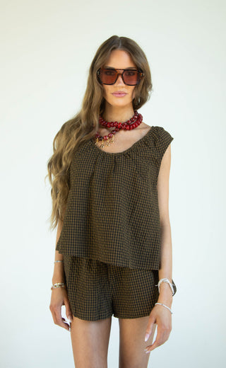 A woman modeling a matching two-piece set consisting of a sleeveless top and shorts in a brown and black gingham pattern, styled with a red beaded necklace and sunglasses against a plain white background.