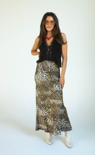 A woman modeling a sleeveless black top and a leopard-print maxi skirt, styled with a red necklace and white pointed boots, against a plain white studio background.
