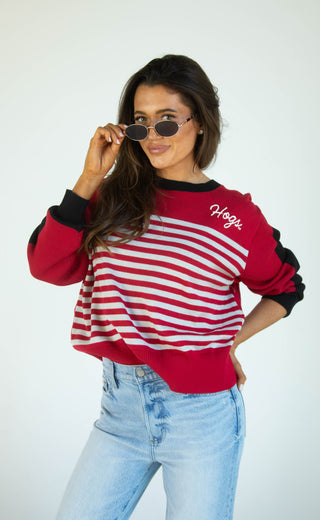 A woman modeling a red and white striped long-sleeve sweatshirt with black trim and the word 'Hogs' embroidered on the chest, paired with light blue jeans against a plain white background.