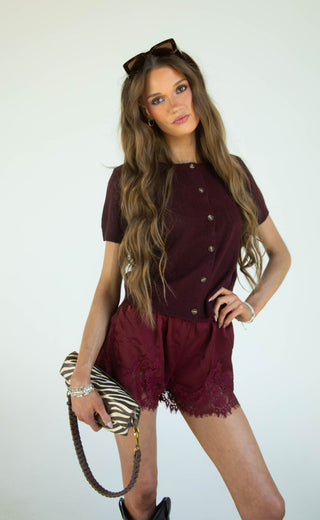 A woman modeling a burgundy short-sleeve button-front top and matching burgundy lace-trimmed shorts, holding a black and white zebra-print clutch against a plain white studio background.
