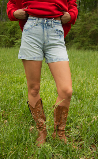Women wearing a red sweater, light blue shorts, and brown cowboy boots standing in a grassy field.