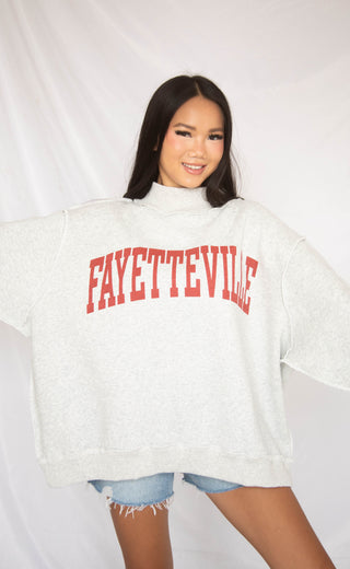 Person wearing a white sweater with 'FAYETTEVILLE' printed in red on a plain background