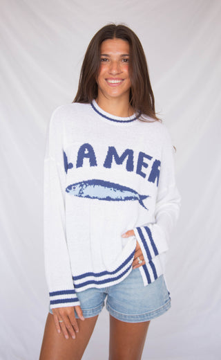 Woman wearing a white sweater with blue text and graphics on a plain background