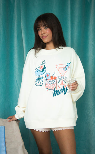 Woman wearing a white sweatshirt with colorful graphics and text, holding a glass against a teal curtain background.