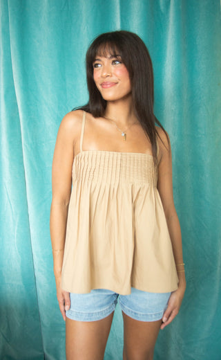 Woman wearing a light taupe pleated tank top and denim shorts against a teal curtain background