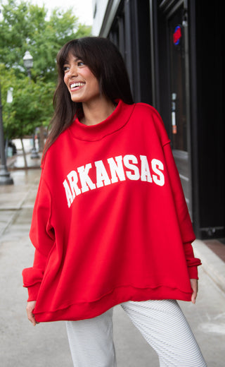 Woman wearing a red 'ARKANSAS ' embroidered mock neck sweatshirt outdoors.