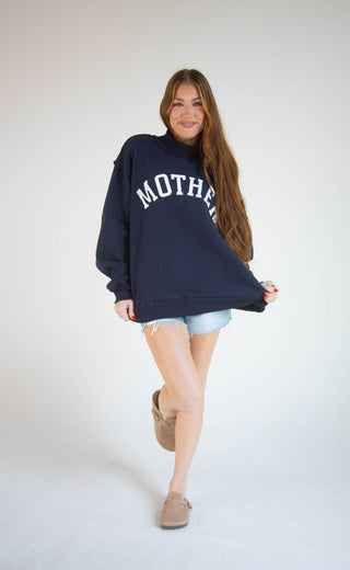 A woman modeling an oversized navy blue sweatshirt with 'MOTHER' printed in white arched letters across the chest, paired with light denim shorts and tan shoes, against a plain white studio background.
