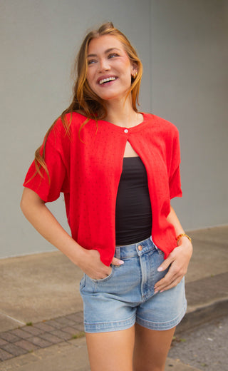 Woman wearing a red sweater cardigan, black top, and denim shorts against a gray background