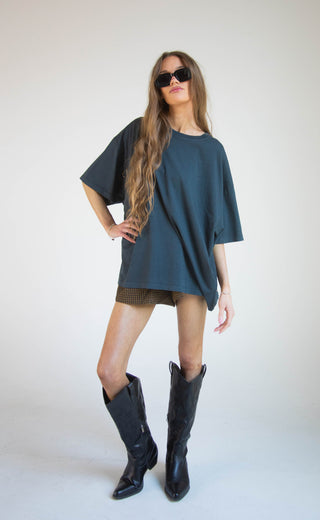 A woman modeling an oversized dark teal blue t-shirt, styled with brown plaid shorts, black cowboy boots, and black sunglasses against a plain light gray studio background.