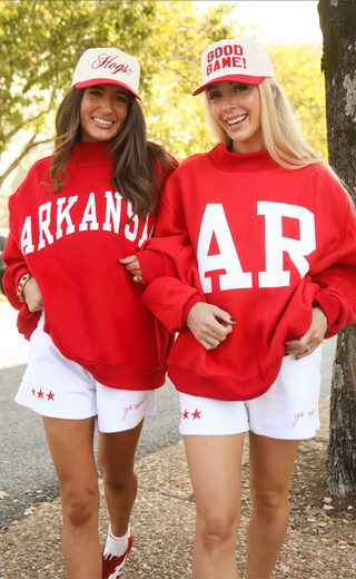 Two women wearing red 'ARKANSAS' sweatshirts and white shorts outdoors.