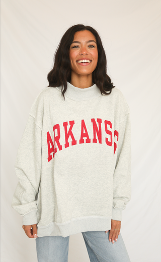 Woman wearing a grey mockneck sweatshirt with 'ARKANSAS' printed in red on a plain background