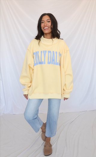 Woman wearing a light yellow mockneck sweatshirt with 'Dilly Dally' text, standing against a white background.
