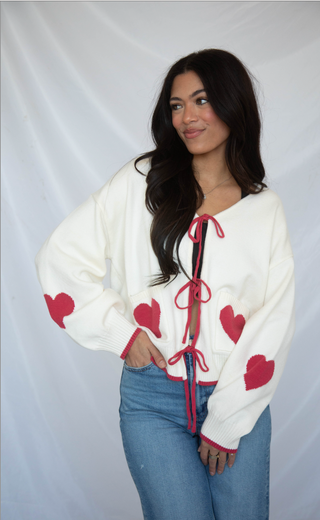 Woman wearing a white sweater with red heart patterns and blue jeans against a plain background