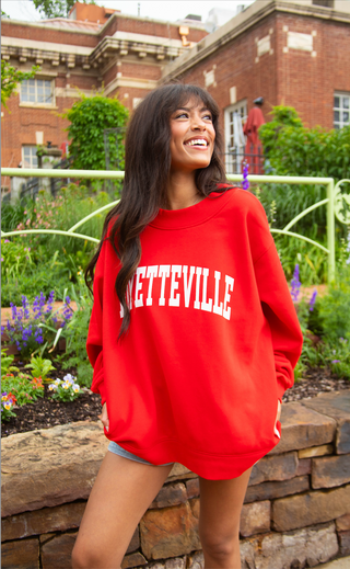 Woman wearing a red sweatshirt with 'FAYETTEVILLE' printed on it, standing outdoors in front of a brick building.