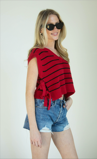 A woman modeling a red and black striped knit top with side ties, paired with denim shorts and black sunglasses, against a plain light background.