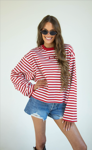 A woman modeling a red and white striped long-sleeve crewneck sweatshirt with a small white embroidered logo on the left chest, paired with denim shorts, against a plain light background.