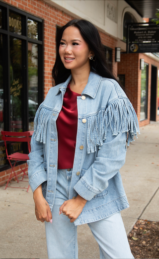 A woman wearing a light-wash denim jacket with fringe details on the front, styled over a dark red satin top and matching light-wash jeans, standing on a sidewalk in front of a brick storefront.