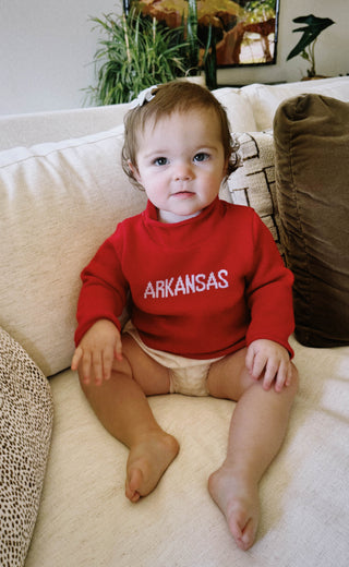 Baby wearing a red 'ARKANSAS ' sweater sitting on a couch.